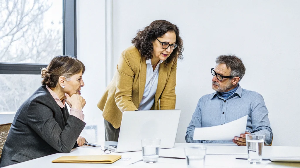 Three executives at a meeting table weighing up a decision, one standing at a laptop while a colleague reviews a printed document